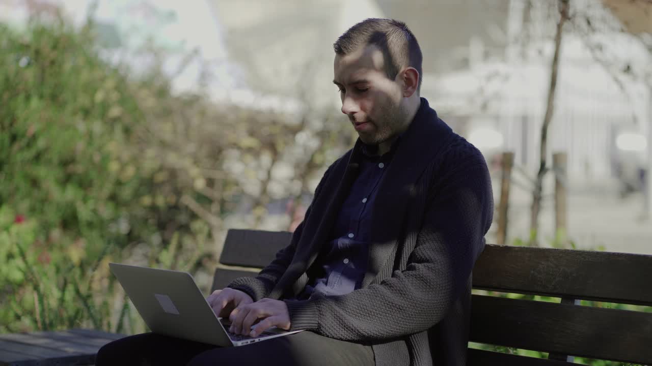 Focused man working with laptop on knees while sitting in park
