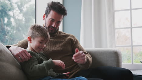 father playing video game with son on mobile phone sitting on sofa at home