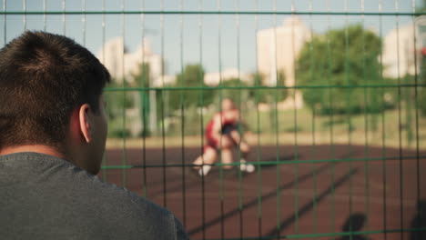 Vista-Trasera-De-Un-Joven-Sentado-Y-Viendo-Una-Sesión-De-Entrenamiento-De-Baloncesto-En-Una-Cancha-De-Baloncesto-Al-Aire-Libre-En-Un-Día-Soleado