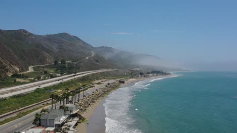 Niedrige-Luftaufnahme-Von-Häusern-Am-Strand-Entlang-Des-Ozeans-Neben-Der-Berühmten-Pacific-Coast-Highway-Route-1-Mit-Wunderschönem,-Nebelverhangenem-Bergblick-An-Einem-Sonnigen-Sommertag