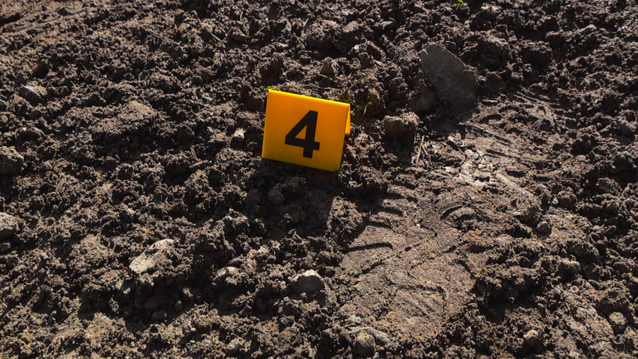 Top down close up view of a yellow crime marker placed next to a footprint left behind by a criminal suspect in mud during a sunny day in a rural area. Details and textures of evidence is visible.