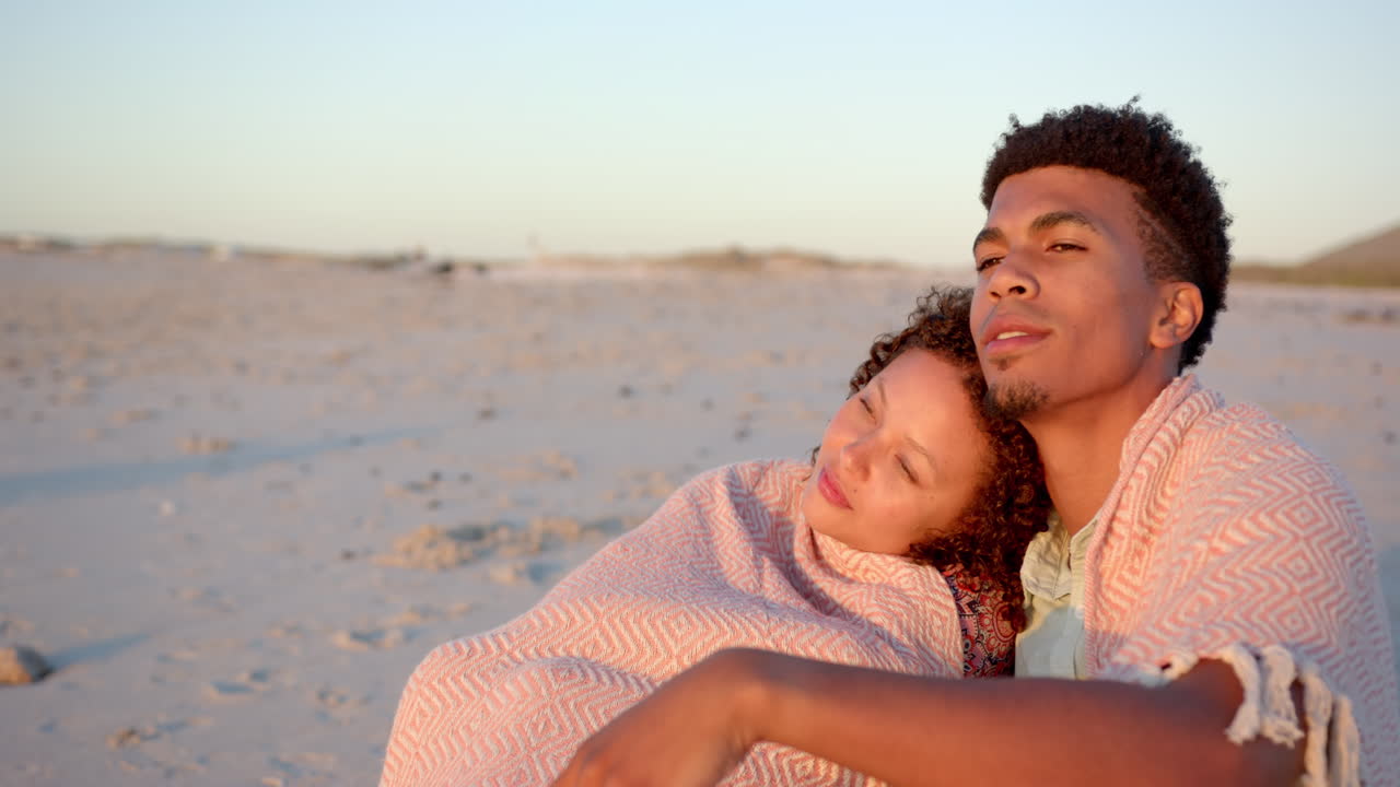 Biracial couple embraces wrapped in a blanket on the beach at sunset with copy space