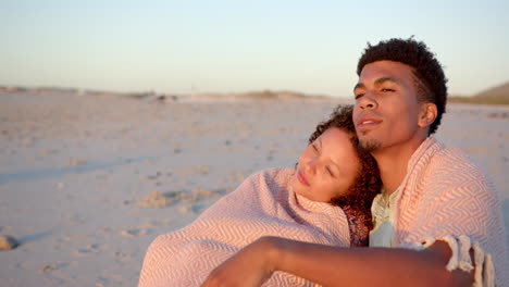 Ein-Biracial-Paar-Umarmt-Sich-In-Eine-Decke-Gehüllt-Am-Strand-Bei-Sonnenuntergang-Mit-Kopierraum