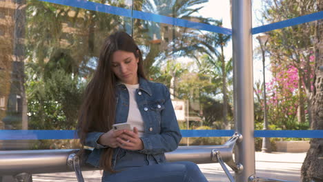 smiling young woman texting on phone sitting at bus stop and then standing up to take the bus