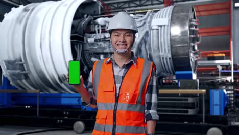 asian male engineer with safety helmet smiling and showing green screen smartphone to the camera while standing with airplane engine maintenance conducted, analytics checking the turbine