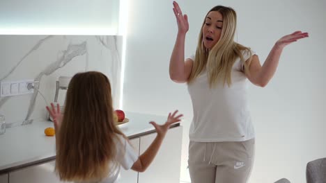 Happy-family-in-the-modern,-white-kitchen.-Mom-and-daughter-in-white-casual-clothes-relaxfully-dance-and-singing.-Mom-is-dancing-with-her-daughter-in-the-kitchen.-Slow-motion