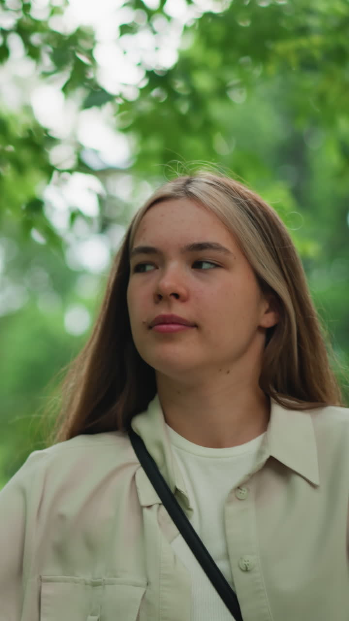 mujer joven con bolsa de cuerpo cruzado negra paseando al aire libre, rodeada de vegetación borrosa y luz solar suave filtrando a través de las hojas, con una vista de fondo de estructuras de construcción y árboles