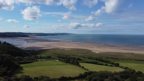 Clip-Cinematográfico-De-Drones-De-4k-Hacia-El-Mar-De-Irlanda-Sobre-Los-Impresionantes-Campos-De-Red-Wharf-Bay-En-Anglesey,-Gales,-Reino-Unido,-Con-El-Pueblo-De-Benllech-Al-Fondo