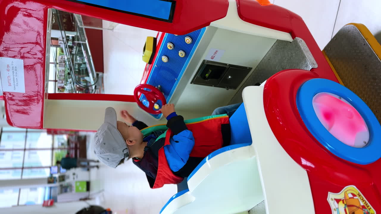 Adorable smiling baby boy in a cap turns the steering wheel of a toy car. Lovely toddler in a firemen car. Vertical screen.