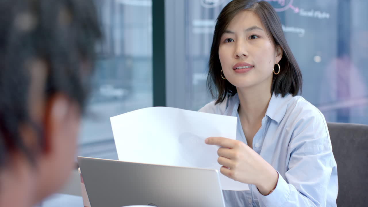 Una donna, tenendo un foglio e usando un laptop, discute il lavoro con un collega in ufficio.