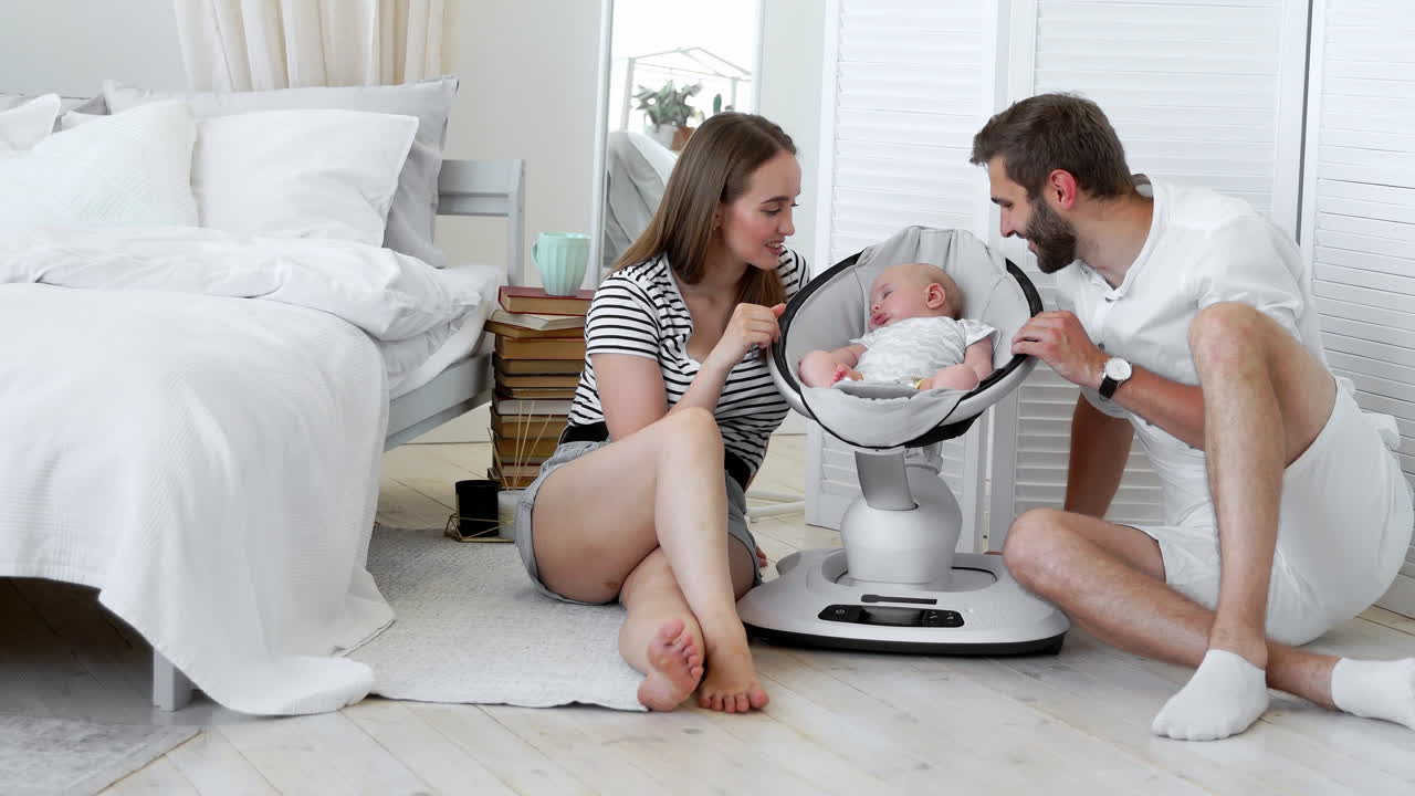 Close - up of happy parents watching their little baby sleep in a rocking infant chair at home in a white apartment. The concept of minimalism