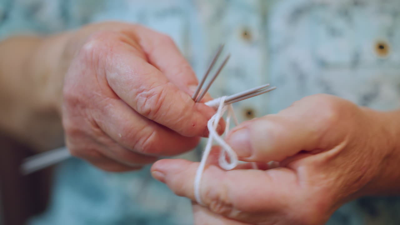 Primer plano macro de los dedos de la abuela tejiendo con varias agujas, manos arrugadas manipulando hilo blanco, puntos y tensión precisos, detalle táctil, artesanía íntima, movimiento repetitivo relajante.