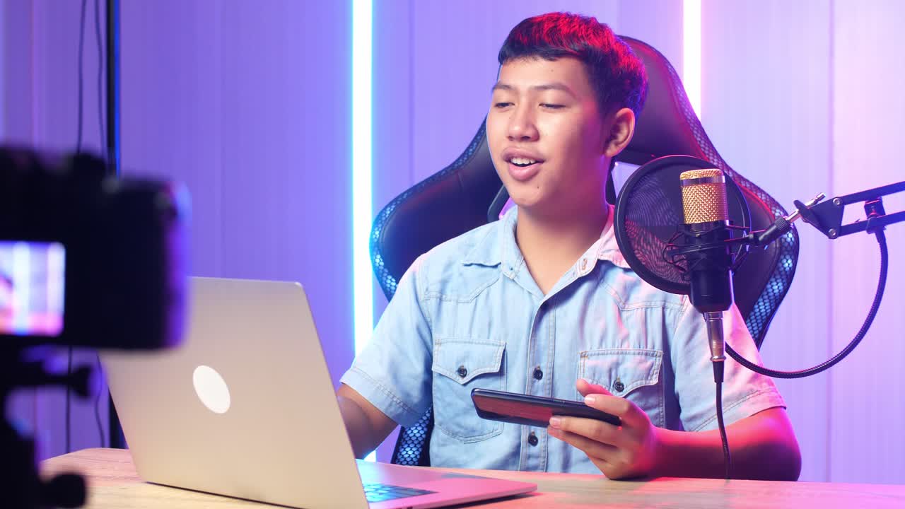 Asian Kid Boy Holding Mobile Phone And Read Comment In Laptop Computer Then Talking To Camera While Live Stream