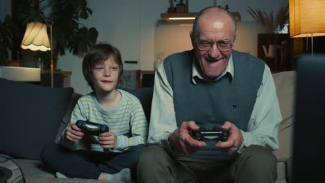 happy senior 60s grandfather having fun playing console game using joysticks with cute teen boy grandson at home on sofa