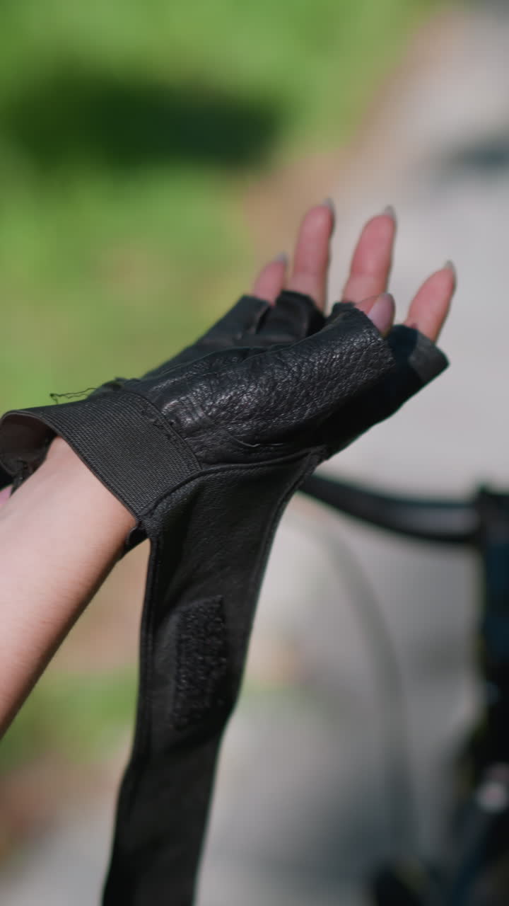 vista trasera de primer plano de un individuo asegurando el guante de motociclista en la mano derecha al lado de la bicicleta, con enfoque en los dedos ajustando la correa, el fondo muestra vegetación borrosa y camino pavimentado