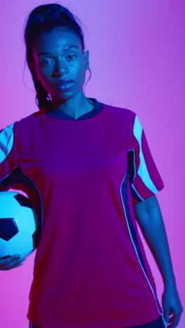 vídeo vertical de primer plano retrato de estudio de una jugadora de fútbol o fútbol femenina con camiseta de equipo sosteniendo la pelota disparada en clave baja contra una iluminación mixta colorida