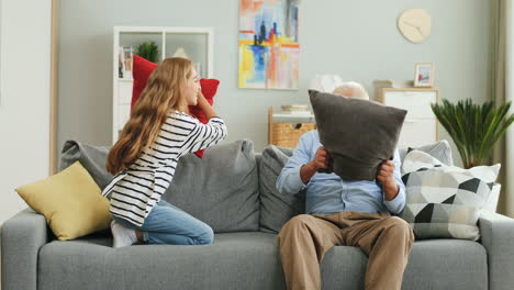 Süßes-Teenager-Mädchen,-Das-Mit-Ihrem-Alten-Großvater-Auf-Dem-Sofa-Im-Gemütlichen-Zimmer-Einen-Kissenkampf-Hat