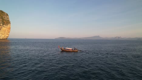 aerial drone approaching a thail longtail boat anchored in the water of krabi thailand during a sunrise tour with a tourist on board and a large limestone rock in the distance