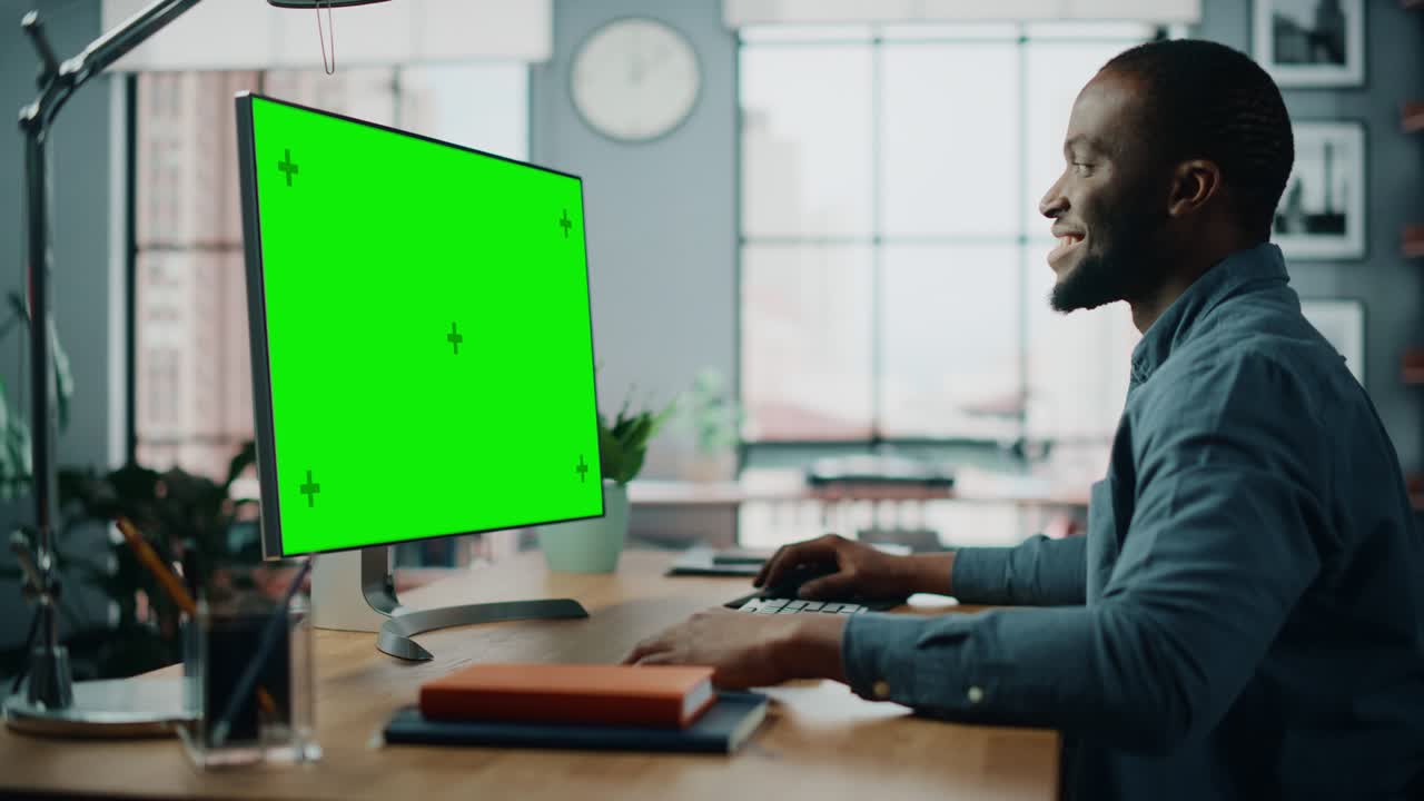 un bellissimo specialista afroamericano nero che lavora su un computer desktop con schermo verde in sala da pranzo a casa. un uomo freelance che chatta con i clienti su internet sui social network.