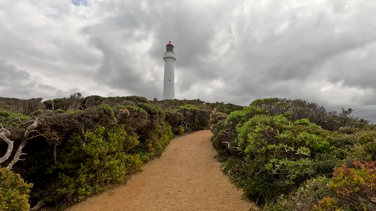 A serene pathway leads to a lighthouse under cloudy skies, surrounded by lush greenery on the Great Ocean Road