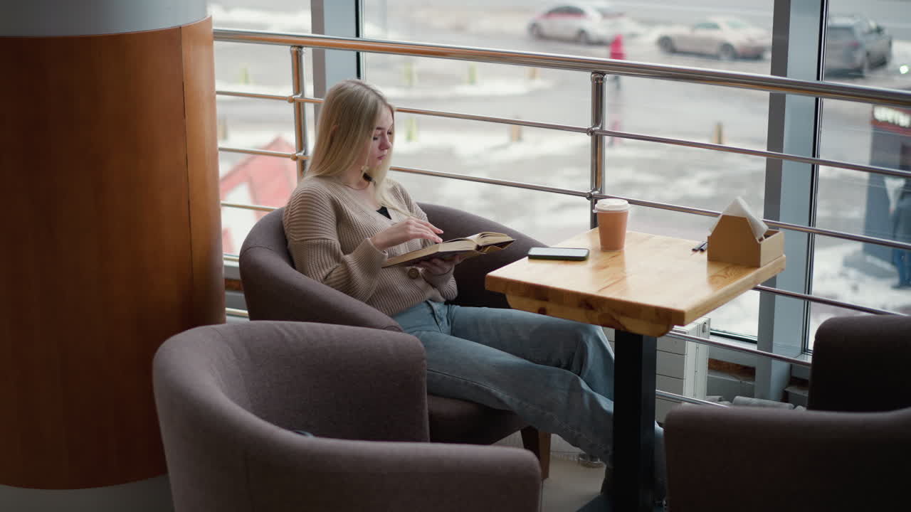 mujer joven sentada pensativa leyendo un libro en un acogedor café, taza de café en la mesa, con una expresión tranquila y pacífica en un entorno urbano moderno con una vista exterior borrosa