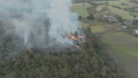 Aerial-drone-shot-in-slow-motion-around-a-mild-forest-fire-on-a-hill-on-a-cloudy-day,-smoke-poised-to-erupt-into-flames