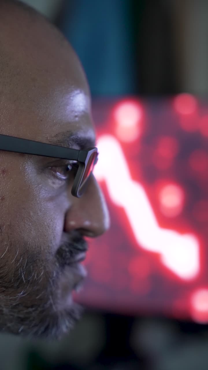 Close up of south asian businessman wearing glasses, observing declining stock market figures on a monitor, shaking head. vertical video