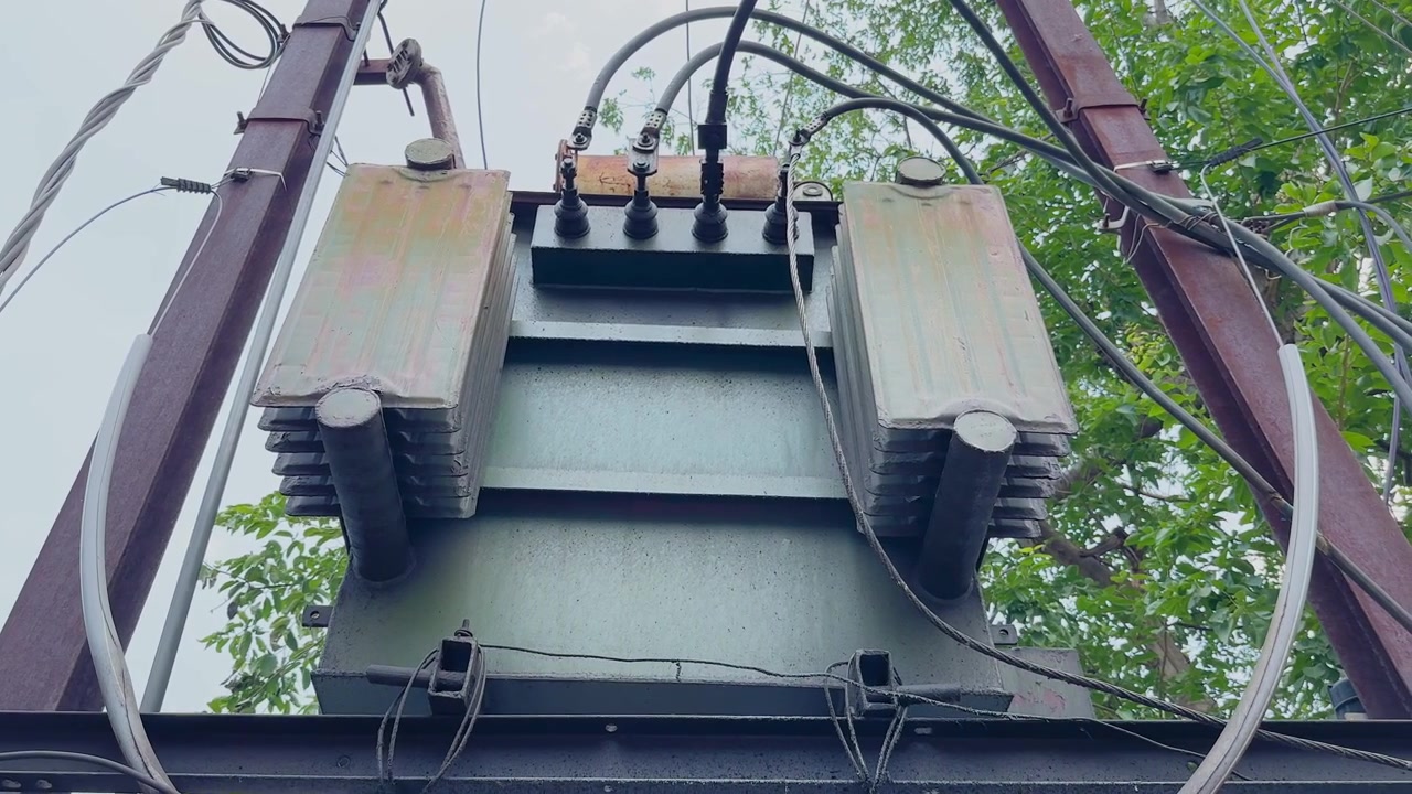 Carrellata da sinistra a destra attraverso un trasformatore di distribuzione elettrica montato su una struttura di servizio, che mostra cavi di alimentazione, alette di raffreddamento e infrastrutture elettriche urbane