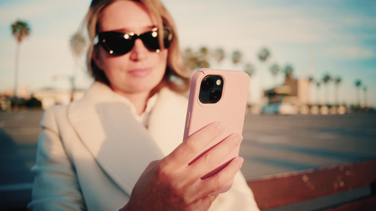 Donna elegante con cappotto bianco guarda il suo smartphone mentre è seduta su una panchina di legno in un parcheggio soleggiato costeggiato da palme