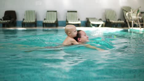 La-Joven-Madre-Está-Nadando-En-La-Piscina-Sosteniendo-A-Su-Pequeño-Y-Lindo-Niño-En-La-Espalda.-Un-Niño-Adorable-Está-Abrazando-A-Su-Madre-Y-Sosteniéndola-Mientras-Nada-En-La-Piscina.-Paseo-A-Cuestas-En-El-Agua