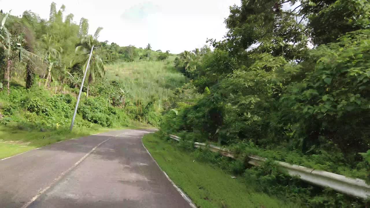 Driving through a long road surrounded by grass and trees. It is a winding road away from the city. The weather is fine and sunny.