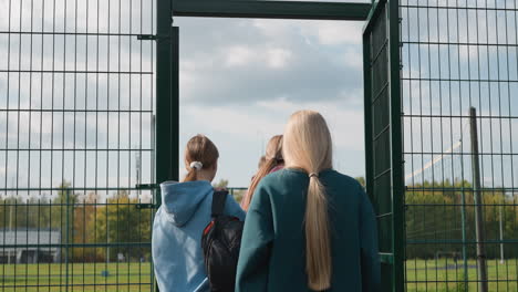 back view of young people entering a volleyball court as coach opens the gate, with a football field and trees visible in the background, emphasizing an active lifestyle