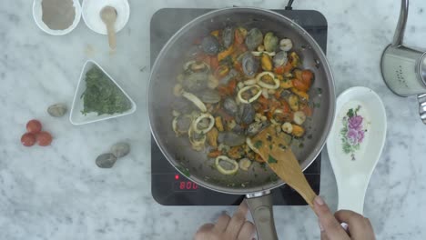 a person stirring seafood with a wooden spoon on a small frying pan, over top shot