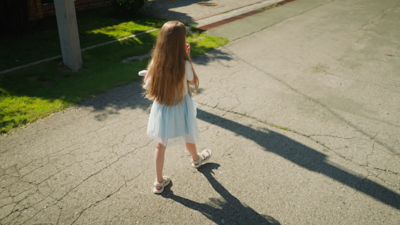 Back view of calm young girl with long hair walking on cracked asphalt road holding something in hand as utility pole casts long shadow across pavement and green lawn edges suburban street scene