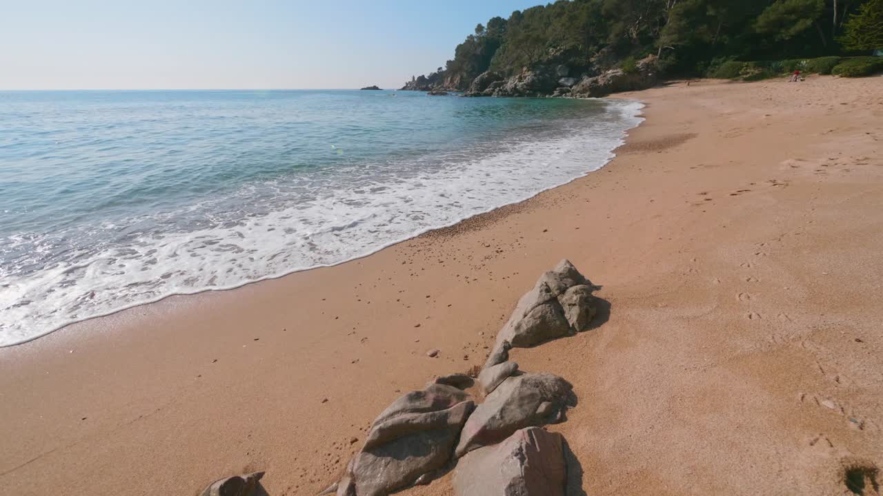 Vlny tyrkysové vody valící se na pláž s růžovým pískem v Lloret de Mar, Španělsko.