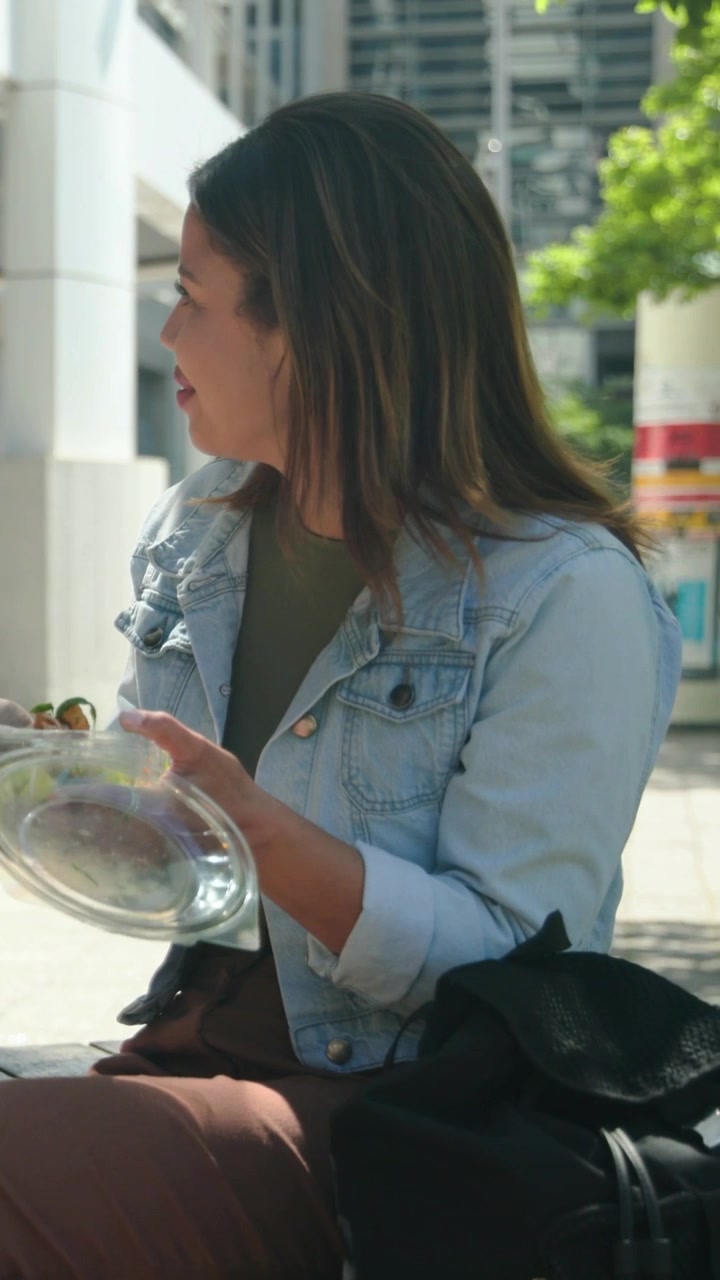Video verticale: Donna in jeans che mangia, guarda in basso e mescola l'insalata per strada durante una pausa, sorridendo.