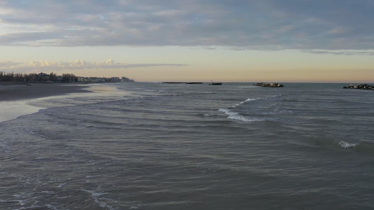 Piccole onde si infrangono sulla riva a Rimini sotto un cielo invernale nuvoloso, con mare calmo, costa distante e frangiflutti al largo in una tranquilla scena costiera.