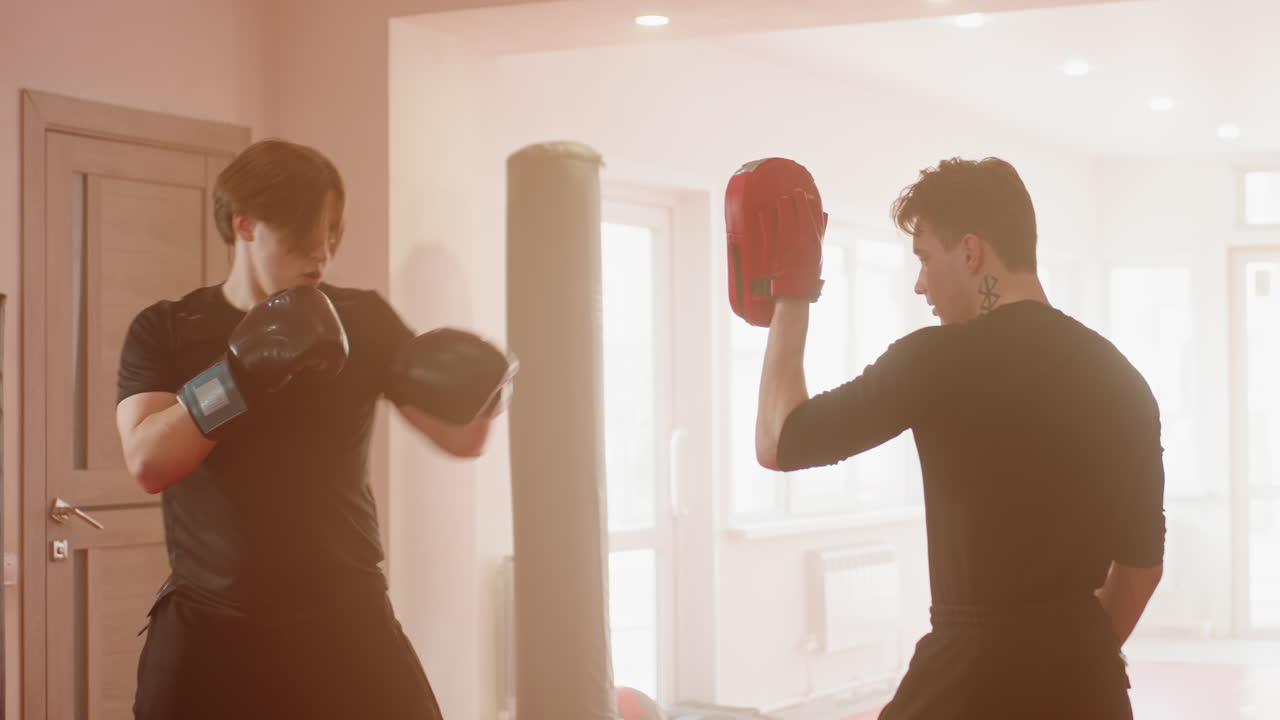 Boxer in black gloves prepares for sparring session with partner holding pads inside gym, showing focus, readiness, stance, determination, discipline, power, energy, and commitment during combat training