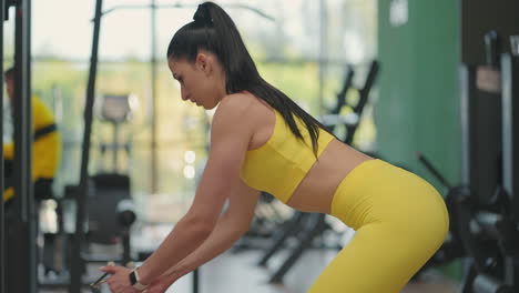 a young hispanic woman in a yellow tracksuit performs an exercise in a crossover pulls a steel rope from below to train her back. a woman trains her back in a gymnasium