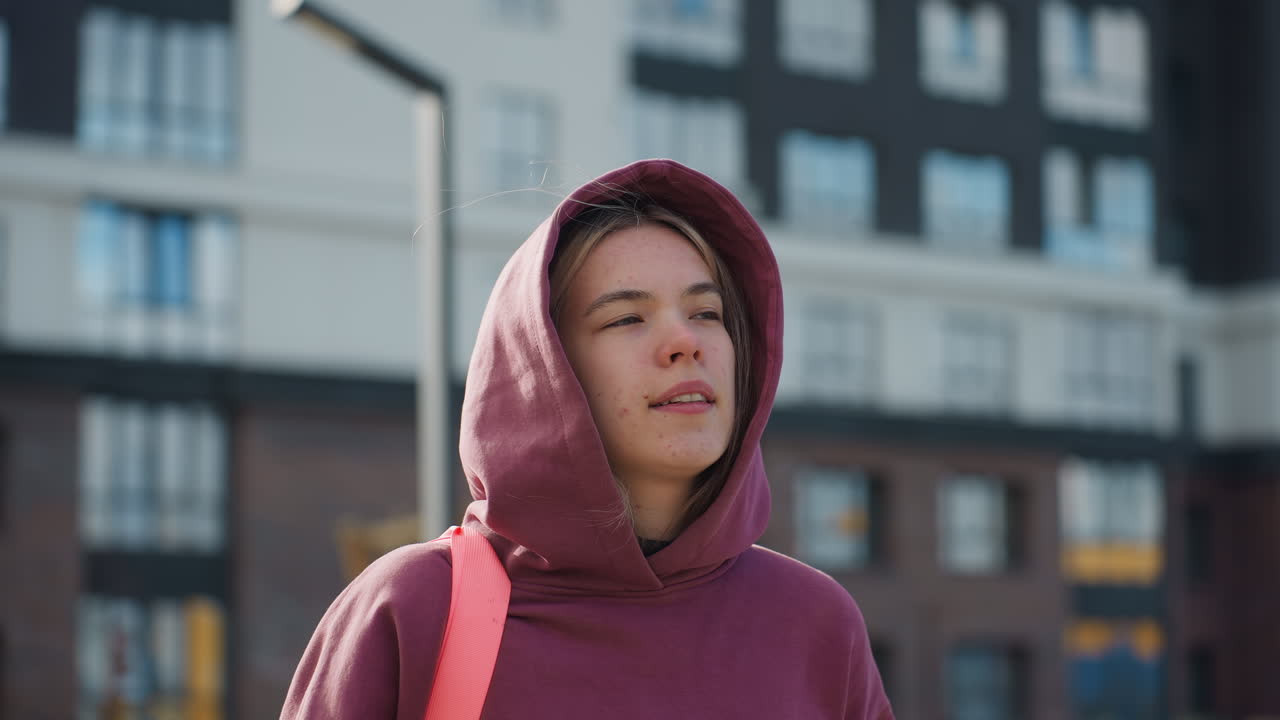 Wellness minded woman hanging bag on shoulders adjusting hoodie while strolling sunny urban street with autumn foliage and modern buildings in background capturing relaxed confidence motion