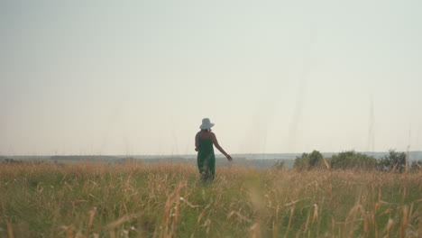 woman wearing wide white hat and green dress walks alone through golden grassy field with camera in hand, bathed in soft natural light and surrounded by countryside horizon under clear