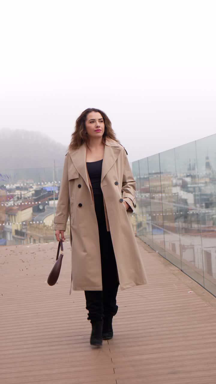 Donna elegante in trench che cammina su una terrazza cittadina.
