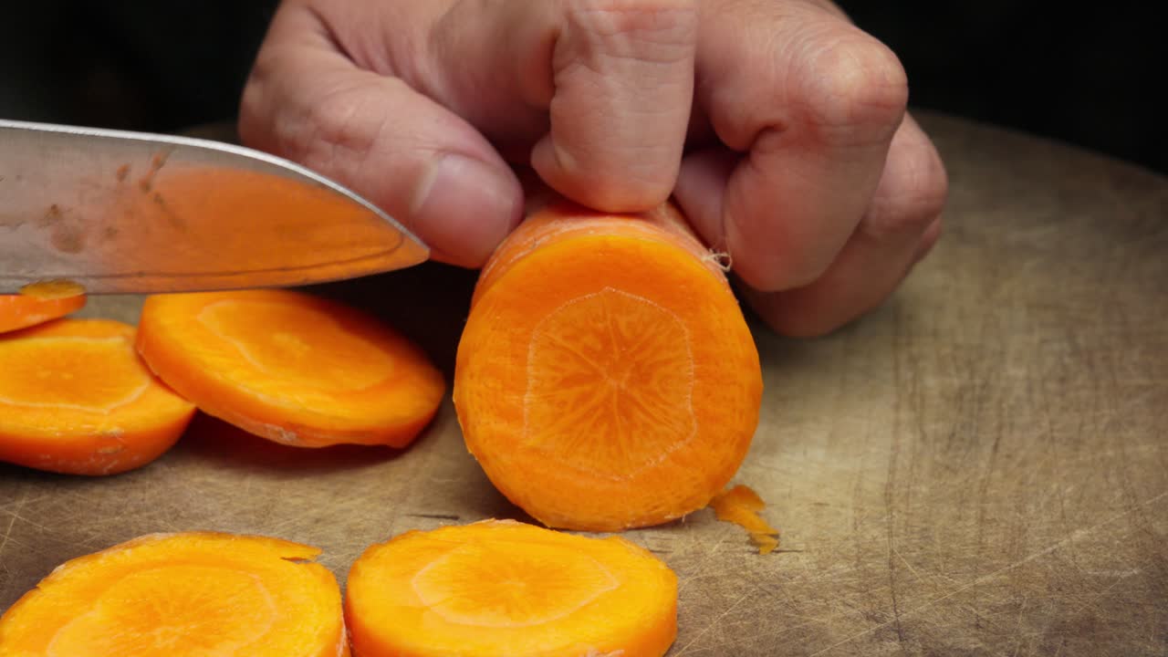 Potret close-up tangan seorang koki menggunakan pisau tajam untuk mengiris wortel segar menjadi irisan bulat di atas talenan kayu. Ideal untuk konten memasak dan nutrisi