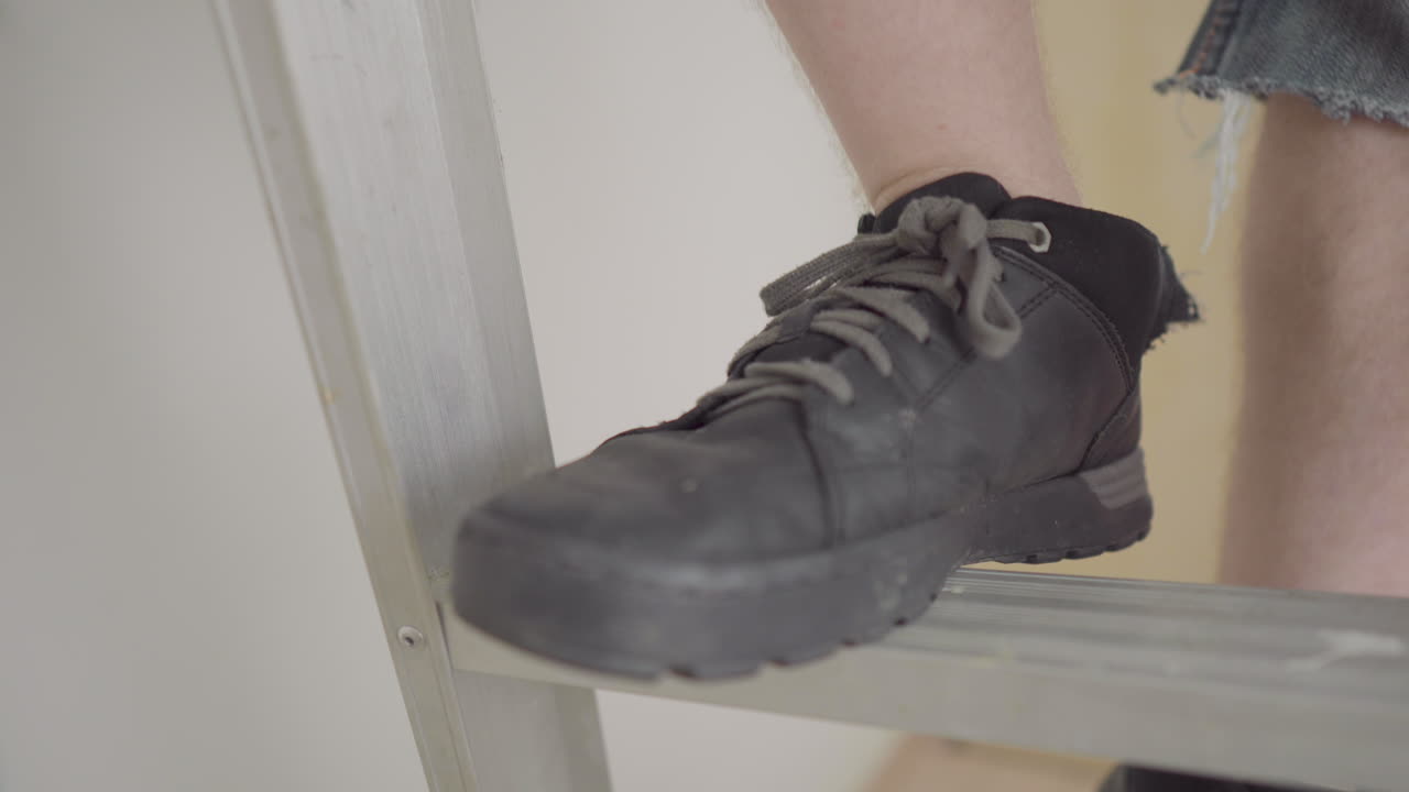 hombre de reparación de viviendas revisando la pared antes de agarrar la herramienta de raspador de enfoque y subir la escalera con sus botas negras dejadas en el marco