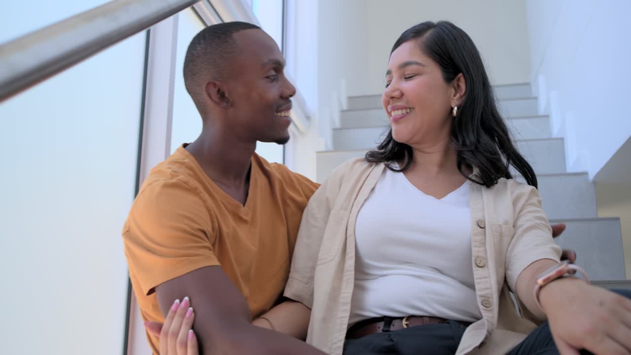 Diverse couple sitting on stairwell by tall window, man wearing orange T-shirt and jeans, smiling