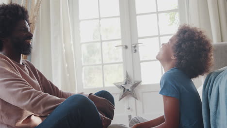 close up of a middle aged black father and his pre teen son sitting cross legged on their living room floor opposite each other, touching heads and smiling, close up