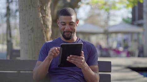 Un-Joven-En-El-Parque-Buscando-Información-En-Una-Tableta,-Riendo