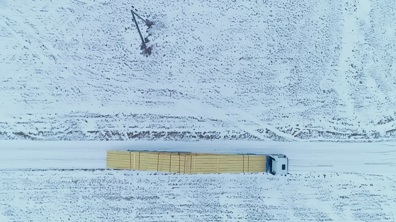 겨울에 눈 덮인 도로에 지붕 건물을 위한 나무 통나무를 운반하는 트럭의 공중 하향식 샷