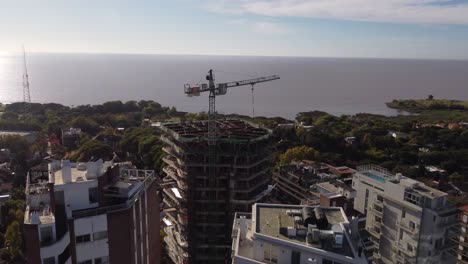 Aerial-view-of-crane-on-top-of-building-roof-under-construction-and-sea-in-background,-Buenos-Aires-in-Argentina