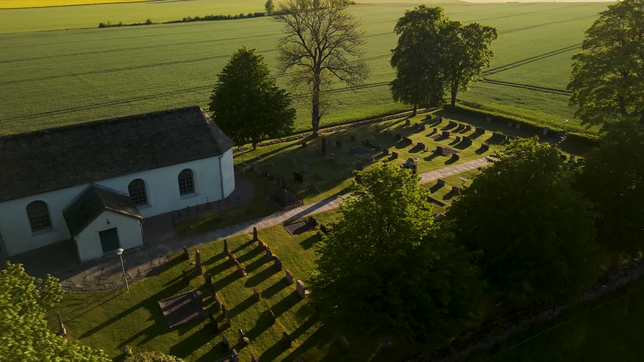 Ripresa parallax con drone che mostra la storica chiesa bianca Järn Church e le lapidi del cimitero durante l'ora d'oro nella campagna svedese con campi agricoli verdi sullo sfondo